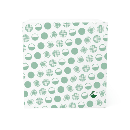 Stack of white sticky notes, the sides of which are printed with a pattern made of circles representing natural elements in white and light teal. One circle is filled in, one is half filled in, one is a starburst, and one is a series of circles in other circles, similar to ripples. On the bottom right corner or each side is The SHE Mark logo in bold teal. 