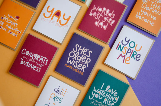 Collection of colorful greeting cards lined up in a diagonal fashion with phrases written in a fun font that say "You're officially over educated and over loved", "Yay you quit your job", "Congrats on your baby business", "World's best girl dad", "She's older and wiser - Happy birthday", and "You Inspire me"