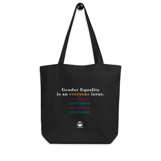 Black tote bag with "Gender equality is an everyone issue" printed on one side. Of that phrase, "gender equality is an" and "issue" are printed in white, and "everyone" is printed in gold. Below the gold "everyone," the word "everyone" is repeated in the following color order: ruby, teal, purple, and green. Below that design is printed the SHE Mark logo in white, with the words "The SHE Mark" in white underneath.