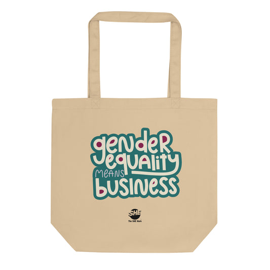 A tan tote bag with the words "gender equality means business" printed in white tet, surrounded by a teal outline. There is ruby color in the open spaces of the letters. Below that text, there is a small black The SHE Mark logo with the words "The SHE Mark printed in black below it.
