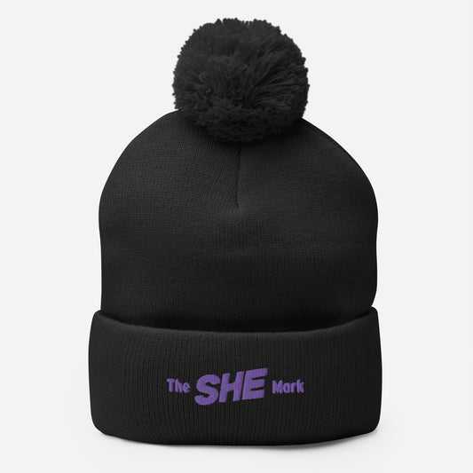 Black knit beanie hat with a same-color pom on top, embroidered on the fold with the words "The SHE Mark" in purple thread.