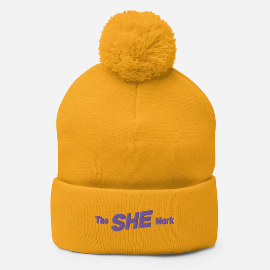 Yellow knit beanie hat with a same-color pom on top, embroidered on the fold with the words "The SHE Mark" in purple thread.