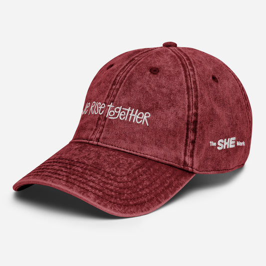 Red dad hat with the phrase "we rise together" embroidered in white on the front. On the left side of the hat embroidered in white is "The SHE Mark." The red of the hat is slightly acid-washed for a vintage look.