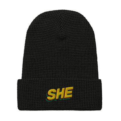 Black waffle knit beanie hat with the word SHE embroidered on the front fold in the color gold, with a green and ruby shadow below it.