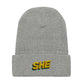 Grey waffle knit beanie hat with the word SHE embroidered on the front fold in the color gold, with a green and ruby shadow below it.